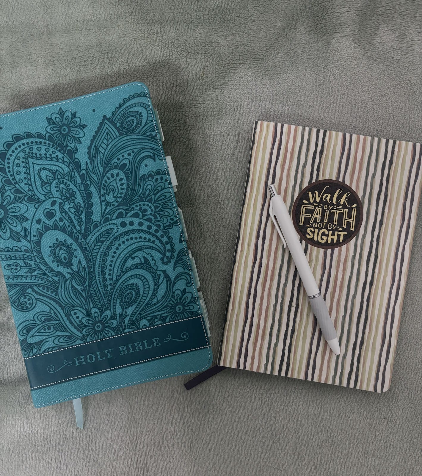 Eine blaugrüne Bibel mit verschlungenen Blumenmustern neben einem gestreiften Tagebuch, auf dessen Einband steht: "Wandle im Glauben, nicht im Schauen." Oben auf dem Tagebuch liegt ein Stift.