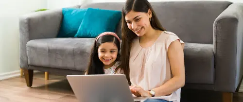 Eine Frau und ein Kind sitzen auf dem Boden, beschäftigt mit einem Laptop in einem gemütlichen Innenraum.