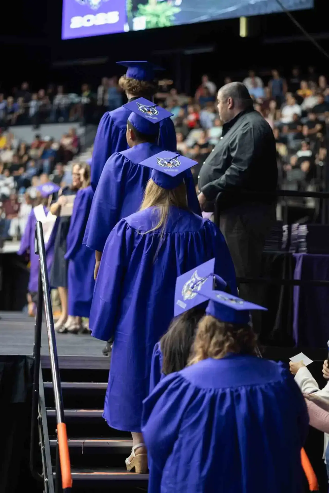 Absolventen in blauen Roben, die mit geschmückten Mützen eine Bühne betreten. Im Hintergrund sieht eine Menschenmenge zu, die eine feierliche Atmosphäre schafft.